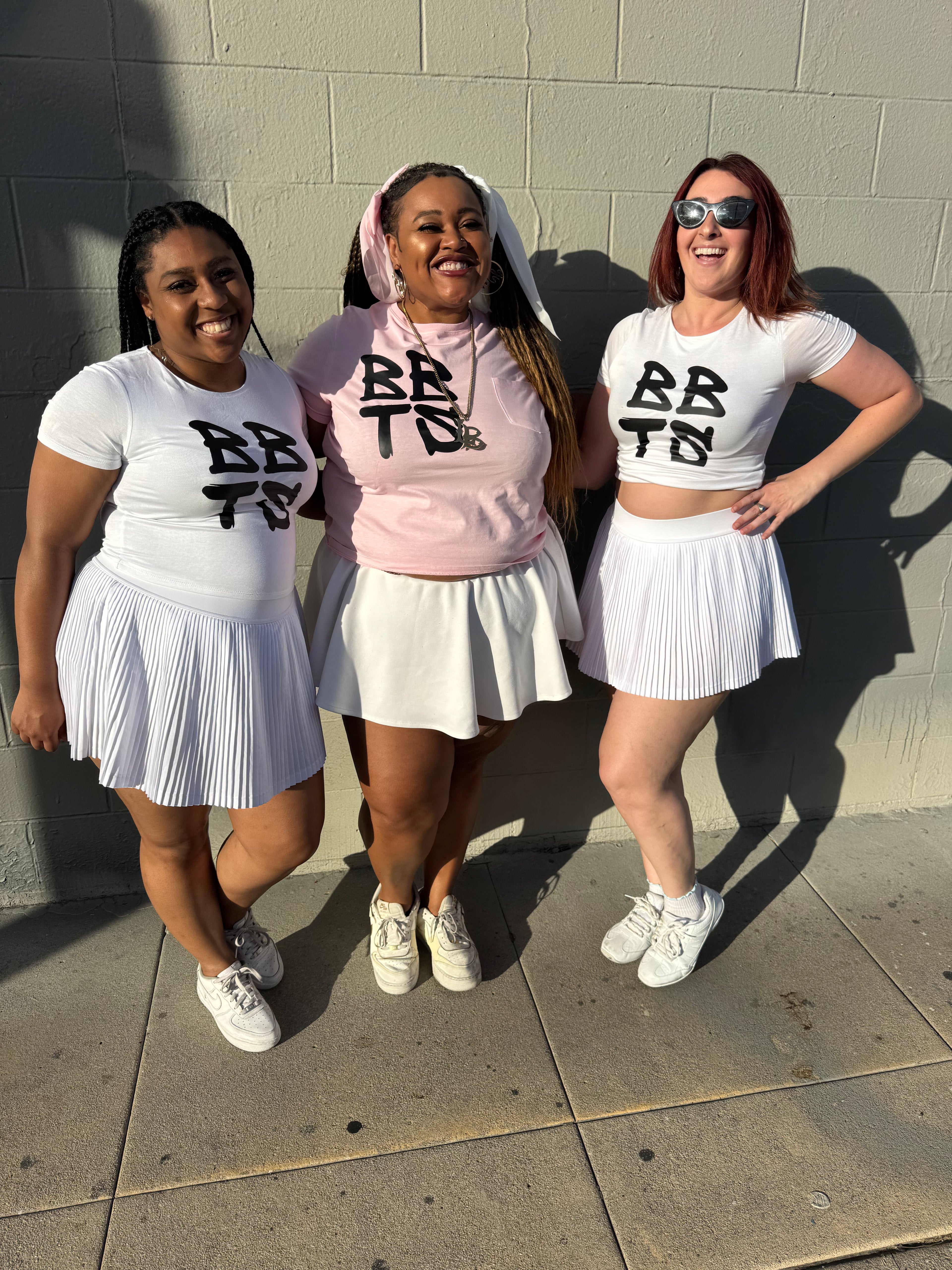 Three women in matching outfits with 'BBT' on a concrete wall background